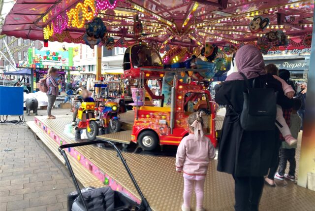 Ein Kinderkarussell mit verschiedenen Fahrzeugen. Davor warten Kinder und ihre Eltern, dass die nächste Fahrt beginnt.