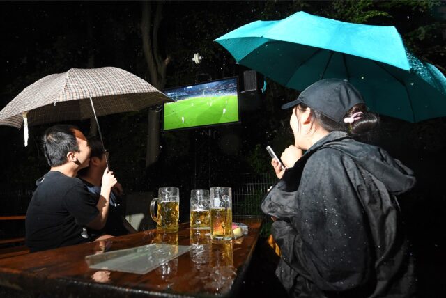 Drei Menschen mit Regenschirmen schauen auf einen Fernseher und gucken ein Fußballspiel. Zwischen ihnen steht ein Tisch mit Bier.