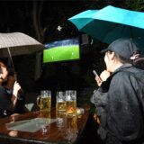 Drei Menschen mit Regenschirmen schauen auf einen Fernseher und gucken ein Fußballspiel. Zwischen ihnen steht ein Tisch mit Bier.