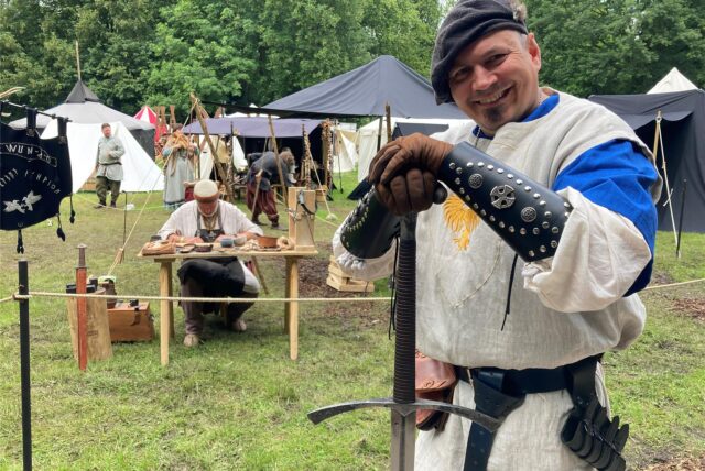 Ein Mann in historischer Kleidung posiert mit einem Schwert auf einem Mittelaltermarkt. Im Hintergrund sind Zelte und weitere Teilnehmer der Veranstaltung zu sehen. (Archiv)
