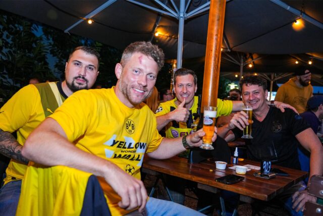 Vier Männer mit BVB-Trikots sitzen an einem Biergarten-Tisch und trinken Bier.