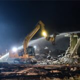 Ein Bagger steht bei Dunkelheit vor einem Schuttberg und teilweise eingestürzten Wand. Aus einem Rohr der Feuerwehr strömt Wasser auf die Ruine.