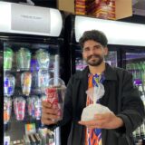 Sores Ekinci steht vor Getränke-Kühlschränken in seinem Vintage Kiosk in Recklinghausen. In der einen Hand hält er einen Plastikbecher, in dem unter anderem eine Dose Coca Cola steckt. In der anderen Hand hält er eine kleine Pastiktüte mit Eiswürfeln.