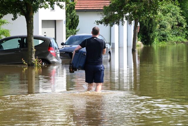 Ein Mann watet mit einem Koffer unter dem Arm durch eine überschwemmte Straße in einem Wohngebiet.