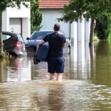 Ein Mann watet mit einem Koffer unter dem Arm durch eine überschwemmte Straße in einem Wohngebiet.