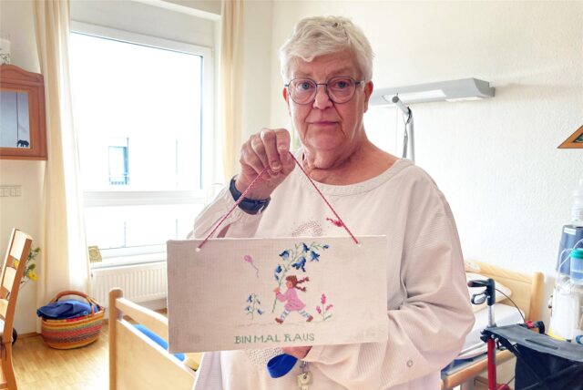 Christel Berg steht inmitten ihres Zimmers im Theodor-Fliedner-Haus in Herten-Westerholt. In der Hand hält sie ein selbst gesticktes Türschild mit dem Schriftzug „Bin mal raus“.