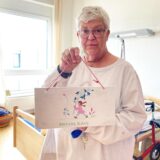 Christel Berg steht inmitten ihres Zimmers im Theodor-Fliedner-Haus in Herten-Westerholt. In der Hand hält sie ein selbst gesticktes Türschild mit dem Schriftzug „Bin mal raus“.