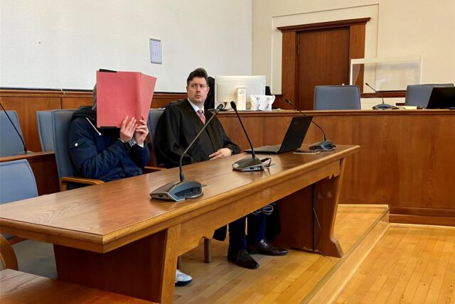 Ein Angeklagter und sein Anwalt sitzen in einem Gerichtssaal. Der Angeklagte hält eine Prozessakte vor sein Gesicht.