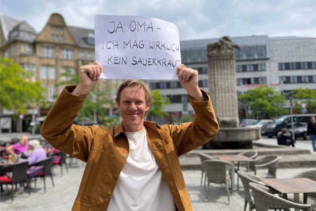 Simon Gosejohann steht auf dem Castroper Marktplatz vor dem Reiterbrunnen und hält einen Zettel mit der Aufschrift „Ja, Oma – ich mag wirklich kein Sauerkraut“ in die Luft.