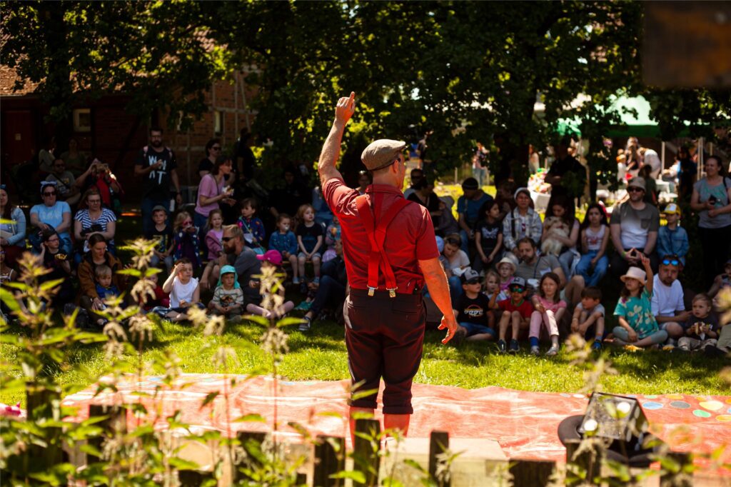 Ein Künstler steht auf einer Wiese, im Hintergrund sind viele Kinder und Erwachsene zu sehen. Ein Künstler steht auf einer Wiese, im Hintergrund sind viele Kinder und Erwachsene zu sehen.