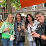 Vier Frauen stehen nebeneinander unter einem mit bunten Segeln geschmückten Baum und halten Gläser in der Hand. Sie lächeln in die Kamera.