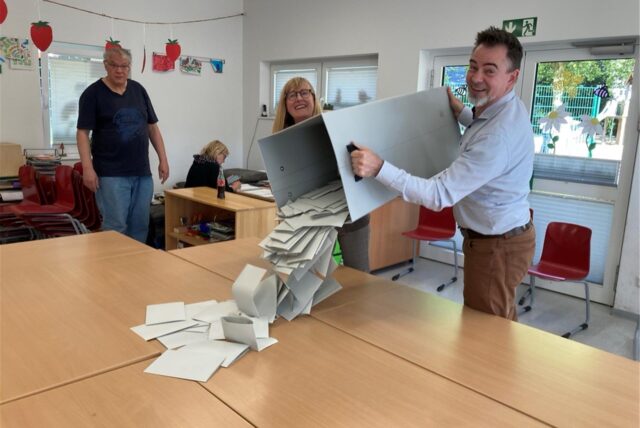 Ein Mann und eine Frau heben eine geöffnete Wahlurne in die Höhe und schütten Stimmzettel auf einen Tisch. Im Hintergrund steht ein Mann und beobachtet den Vorgang.