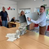 Ein Mann und eine Frau heben eine geöffnete Wahlurne in die Höhe und schütten Stimmzettel auf einen Tisch. Im Hintergrund steht ein Mann und beobachtet den Vorgang.