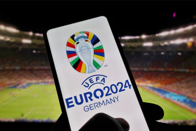 Eine Hand hält ein Smartphone mit dem Logo der EM 2024. Im Hintergrund ist ein Fußballstadion zu sehen.