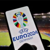 Eine Hand hält ein Smartphone mit dem Logo der EM 2024. Im Hintergrund ist ein Fußballstadion zu sehen.