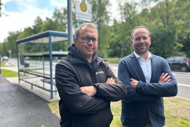 Christoph Brinks, Standortleiter von Arvato, und Thomas Gries, Standortleiter vom DHL-Verteilzentrum in Dorsten stehen mit verschränkten Armen vor einem Bushaktestellen-Häuschen.