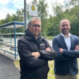 Christoph Brinks, Standortleiter von Arvato, und Thomas Gries, Standortleiter vom DHL-Verteilzentrum in Dorsten stehen mit verschränkten Armen vor einem Bushaktestellen-Häuschen.