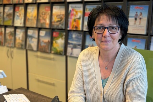Eine Frau sitzt an ihrem Schreibtisch. Sie hat dunkle, kurze Haare und eine Brille. Im Hintergrund ist ein Regal mit Reisekatalogen.