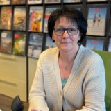 Eine Frau sitzt an ihrem Schreibtisch. Sie hat dunkle, kurze Haare und eine Brille. Im Hintergrund ist ein Regal mit Reisekatalogen.