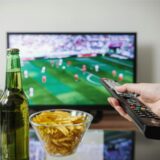 Eine Person hält eine Fernbedienung über einem Tisch, auf dem zwei Flaschen Bier und eine Schale mit Chips steht. Im Hintergrund läuft ein Fußballspiel auf einem Fernseher.