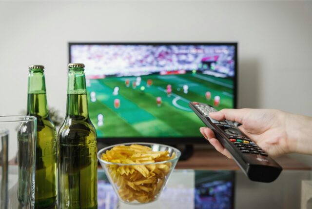 Eine Person hält eine Fernbedienung über einem Tisch, auf dem zwei Flaschen Bier und eine Schale mit Chips steht. Im Hintergrund läuft ein Fußballspiel auf einem Fernseher.