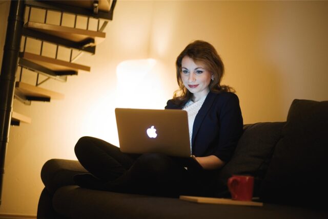 Eine Frau sitzt auf ihrer Couch am Macbook und hat rechts von sich eine Getränketasse stehen.