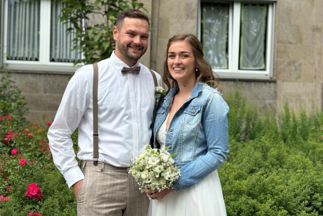 Ein Mann im Anzug und eine Frau im Brautkleid und Jeansjacke lächeln. Sie hält einen Brautstrauß in der Hand.