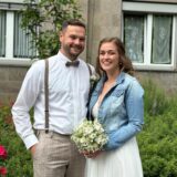 Ein Mann im Anzug und eine Frau im Brautkleid und Jeansjacke lächeln. Sie hält einen Brautstrauß in der Hand.