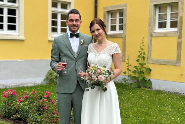 Ein Mann im Anzug und eine Frau im Brautkleid stehen vor einem gelben Gebäude im Garten. Sie halten beide ein Sektglas. Die Braut hat auch einen Strauß.