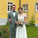Ein Mann im Anzug und eine Frau im Brautkleid stehen vor einem gelben Gebäude im Garten. Sie halten beide ein Sektglas. Die Braut hat auch einen Strauß.