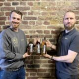 Andree Stryewski (links) und Björn Nessmann stehen vor einer Backsteinwand und halten drei Flaschen mit ihrem eigenen Whisky in der Hand, der in Räumen der Hertener Zeche Schlägel und Eisen drei Jahre lang lagerte.