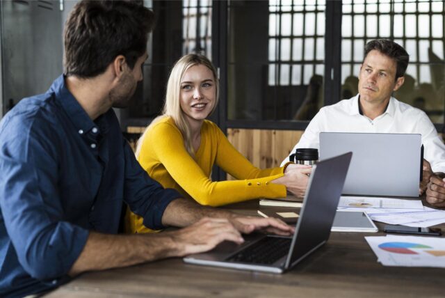Zwei Männer und eine Frau diskutieren an einem Tisch mit Laptops über Business-Entscheidungen.