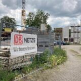 An einem Bauzaun an der Kaiserstraße in Herten hängt ein Banner der Deutschen Bahn mit dem Aufdruck „Wir bauen Brücken“. Im hinteren Teil des Bildes ist die halbfertige Konstruktion einer Brücke zu sehen.