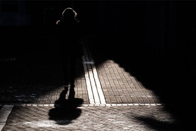 Eine Frau geht über eine Straße, die halb im Schatten liegt.