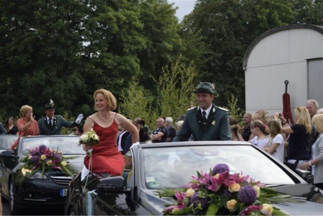 Ein Mann in Uniform und eine Frau im Abendkleid sitzen erhöht in einem Cabrio.