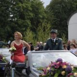 Ein Mann in Uniform und eine Frau im Abendkleid sitzen erhöht in einem Cabrio.