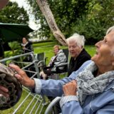 Eine ältere Dame sitzt rechts im Bild und lacht froh. Im Hintergrund sind Schafe und Schildkröten in einem Gehege zu erkennen.