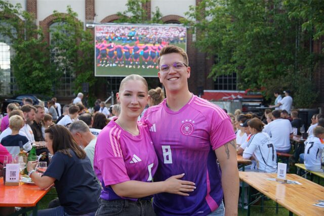 Ein Pärchen steht in pinkfarbenen Fußballtrikots vor einer Leinwand. Im Hintergrund sitzen Menschen an Bierzeltgarnituren.