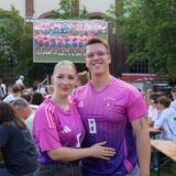 Ein Pärchen steht in pinkfarbenen Fußballtrikots vor einer Leinwand. Im Hintergrund sitzen Menschen an Bierzeltgarnituren.