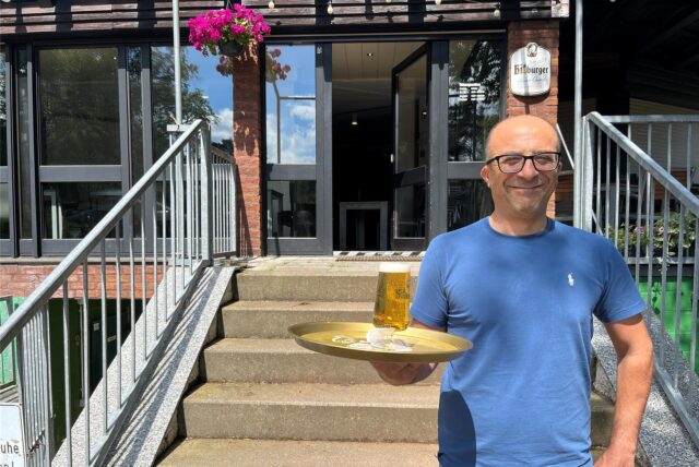 Mann im blauen T-Shirt hält ein Tablett mit einem Glas Bier und steht vor dem Lokal.