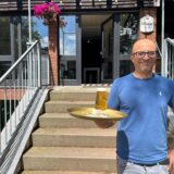 Mann im blauen T-Shirt hält ein Tablett mit einem Glas Bier und steht vor dem Lokal.