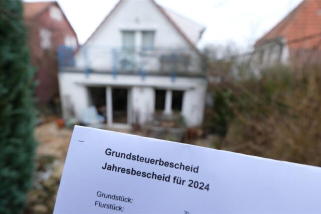 Ein Grundsteuerbescheid für 2024 wird vor einem Privatgrundstück hochgehalten.