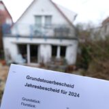 Ein Grundsteuerbescheid für 2024 wird vor einem Privatgrundstück hochgehalten.
