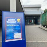 Auf einem Klappaufsteller vor der Lidl-Filiale an der Sachsenstraße in Recklinghausen steht Wir bauen für dich um. Im Hintergrund sind der Eingangsbereich des Discounters und ein grüner Container zu sehen.