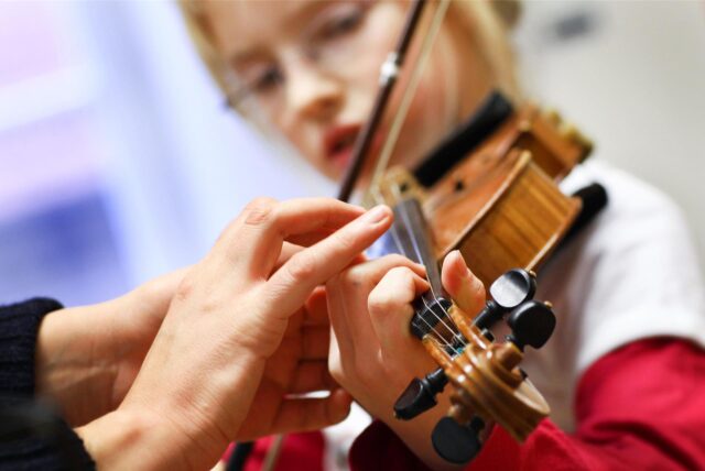 Eine Musiklehrerin korrigiert die Hand einer Schülerin beim Geigenspiel.