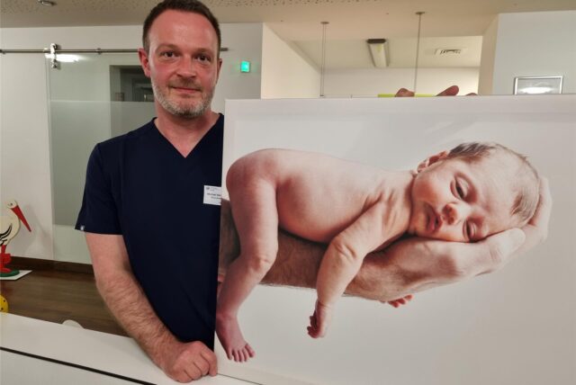 Michael Melchisedech steht im St.-Vincenz-Krankenhaus in Datteln neben einem Bild, auf dem ein Baby auf der Hand eines Mannes liegt.