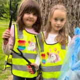 Amelie (l.) und Sofie (r.) stehen auf einer Wiese in Castroper Stadtgarten, tragen Warnwesten und halten einen Müllsack.