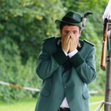 Ein Mann in Schützenuniform steht neben einem Gewehr und schlägt die Hände vors Gesicht,