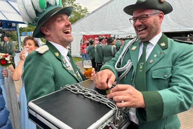 Zwei Schützen lachen, einer öffnet eine Eisenkette, die um einen Koffer gewickelt wurde.