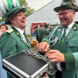 Zwei Schützen lachen, einer öffnet eine Eisenkette, die um einen Koffer gewickelt wurde.